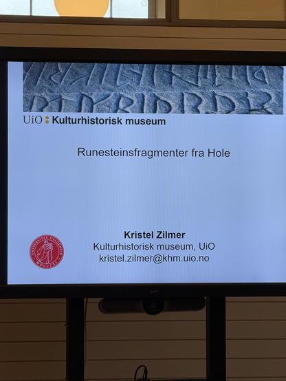 Photo of a screen showing the first slide of a presentation. At the top is a picture of runes carved in stone. The text in the middle says “Runesteinsfragmenter fra Hole”. The name at the bottom is “Kristel Zilmer”