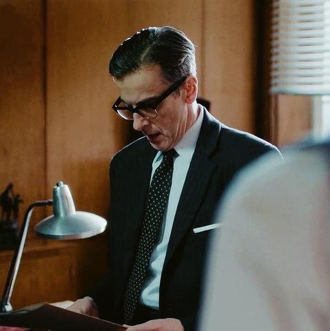 Randall Brown from The Hour is looking down at a folder. He's wearing dark-rimmed glasses, a nice suit, and has his hair slicked back.