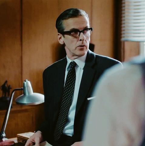 Randall Brown from The Hour is looking to the side at someone as they speak. The snapshot was only taken moments after the first one. Again, he's wearing dark-rimmed glasses, a nice suit, and has his hair slicked back.