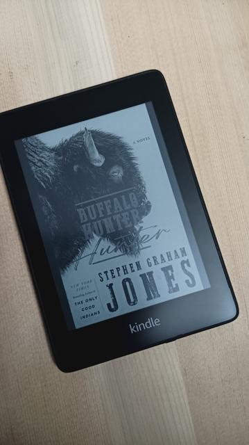 Kindle device on a light brown wooden background showing the cover of the book 'The Buffalo Hunter Hunter'.