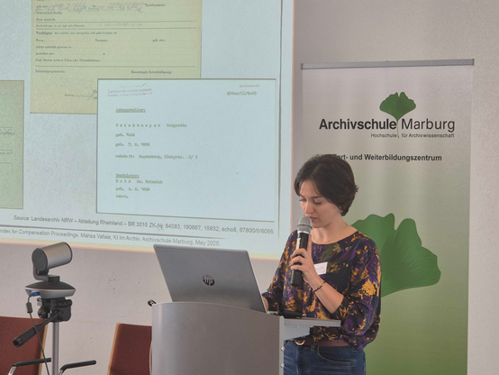 Mahsa Vafaie is presenting at the 29. Archivwissenschaftliches Kollowuium. She is standing behind a desk with an open laptop in front. Behind at right there is part of a slide displayed showing different index cards. At left there is a roll-up banner of Archivschule Marburg.
