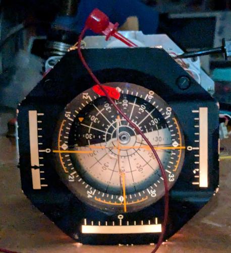 An indicator with a rotating ball in the middle. The top half of the ball is black and the bottom is white. The ball has various lines and angle markings on it. Three yellow needles project across the ball to indicate the error in each axis. Three scales with needle pointers show the rotation rate in the three axes. The scales are illuminated. Two power wires run to clips attached to the interior of the FDAI, which has its cover removed.