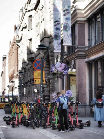 A city street scene featuring e-scooters parked along the sidewalk. A person is examining their phone near the scooters, while another individual stands further down the street. Nearby, bicycles are also parked, and there are decorative banners and flowers along the buildings.