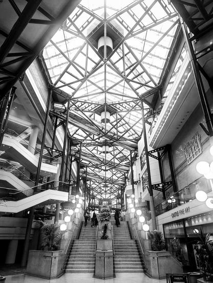 Looking up at a glass roof inside a large building. The roof is covered in supporting beams. The roof is several floors high, with the different levels visible on either side of the walkway. At the bottom of the picture are a double set of stairs leading along the walkway.