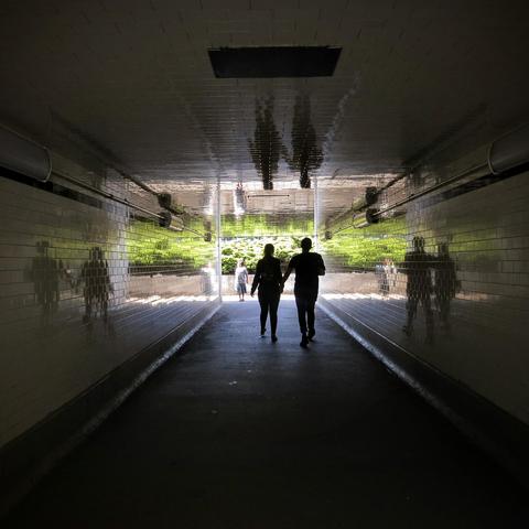 View down a white tiled tunnel towards the end filled with light and greenery. Two outlined figures are walking ahead, and their shapes are reflected in the walls on either side and the ceiling.