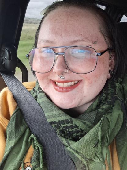 Trans/nonbinary person (they/them) with facial piercings, glasses, and minimal makeup, wearing a yellow SpongeBob sweater and green Palestinian kiffiyeh in the van on the way to an OT appointment.