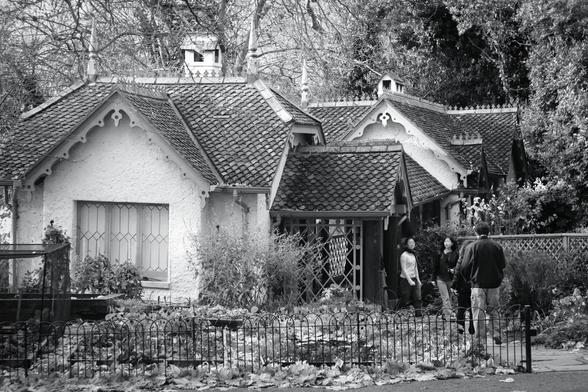 Black and white. Four people, one man and three women, are standing or walking in the front garden of Duck Island Cottage in St James's Park. It is a Swiss chalet-style cottage with two small buildings connected by a covered walkway that crosses a bridge, with tiled roofs, decorated gables, several small spires, and a tall white chimney stack topped with what looks like a small bird house. Seen in colour, it looks somewhat like a gingerbread cottage. It is often topped off by a grey heron enjoying the view over the lake behind the cottage.