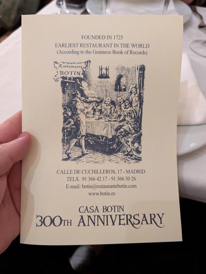 The image shows a card held by a hand, featuring information about Casa Botín, the earliest restaurant in the world according to the Guinness Book of Records, founded in 1725. The card is cream-colored with black text and illustrations. At the top, it states "FOUNDED IN 1725 EARLIEST RESTAURANT IN THE WORLD (According to the Guinness Book of Records)." Below this, there is an illustration of a dining scene with people seated around a table, and a server in an apron. The address "CALLE DE CUCHILLEROS, 17 - MADRID" is printed, along with two telephone numbers: "TELS. 91 366 42 17 - 91 366 30 26." An email address "botin[@]restaurantebotin.com" and a website "www.botin.es" are also provided. At the bottom, it reads "CASA BOTIN 300TH ANNIVERSARY." The card is set against a table with a white tablecloth, a plate, and a glass, suggesting a dining setting.

Provided by @altbot, generated privately and locally using Ovis2-8B