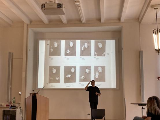 Large seminar room with a teacher dressed in black at the front signing. Displayed on the screen is a page from the first attempt to collect signs that would be internationally understood.