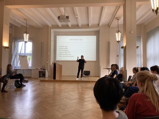 Large seminar room with nice wooden flooring with participants sat on chairs in a circle. On the board, there are some bullet points about International Sign.