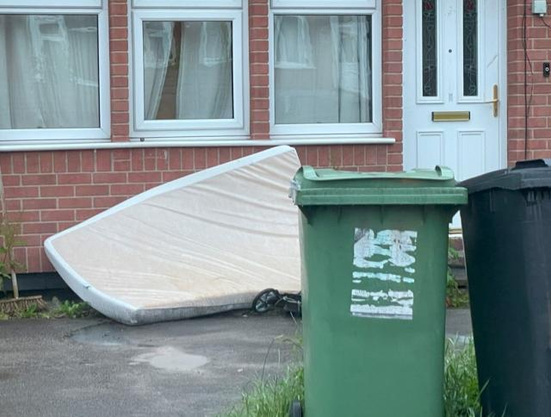 a mattress against a house and there are two wheelie bins in the foreground