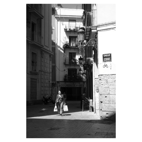 This is a portrait format black and white image of a woman carrying shopping bags walking through a patch of sunlight on a street otherwise in shadow. Taken in Valencia, Spain, with a Sony RX100iii.