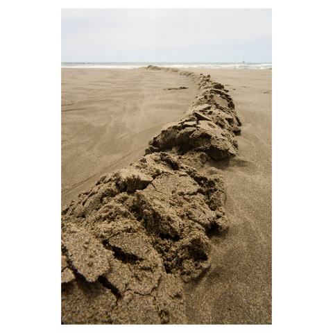Wide angle image taken on a beach. The camera is close to the ground, focused on a heap of sand forming a long curve that runs down the beach to the water. The sea and a sailboat are visible at the top of the frame. The sky is cloudy.