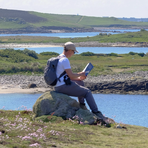 A man in grey trousersand white t-shirt sits on a rock consulting a guidebook. He wears a beige cap and a grey rucksack. A pair of binoculars hang by his side. The biew behind is a mix of grassy islands and blue ocean water