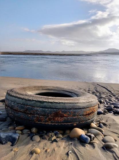 In front, a large, old car tyre lies between stones on a sandy beach. The blue-grey water behind it lies calmly in the large bay. A sandbank can be seen in the background, then on the horizon the opposite shore of the bay with the Ox Mountains, which lie hazy in the sun under a slightly cloudy sky.
