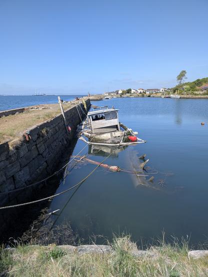 The image depicts a serene coastal scene with a calm body of water reflecting the clear blue sky. In the foreground, a small white boat with a red stripe is anchored near the shore, tethered by a rope to a buoy. To the left, a partially submerged wooden structure with a red object, possibly a buoy or a piece of debris, is visible. The water is clear, with some seaweed and rocks near the shore.

In the background, a line of trees and a stone structure, possibly ruins or remnants of a historical building, are visible along the water's edge. Several houses with red roofs are scattered along the shoreline, adding to the tranquil atmosphere. The sky is mostly clear with a few scattered clouds, enhancing the peaceful ambiance of the scene