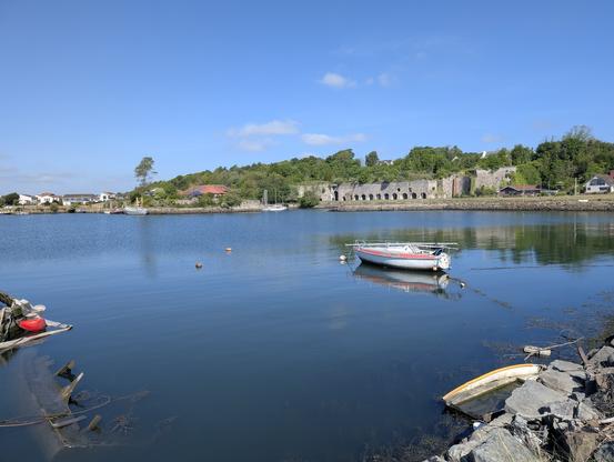 The image depicts a serene coastal scene with a calm body of water reflecting the clear blue sky. In the foreground, a small white boat with a red stripe is anchored near the shore, tethered by a rope to a buoy. To the left, a partially submerged wooden structure with a red object, possibly a buoy or a piece of debris, is visible. The water is clear, with some seaweed and rocks near the shore.

In the background, a line of trees and a stone structure, possibly ruins or remnants of a historical building, are visible along the water's edge. Several houses with red roofs are scattered along the shoreline, adding to the tranquil atmosphere. The sky is mostly clear with a few scattered clouds, enhancing the peaceful ambiance of the scene