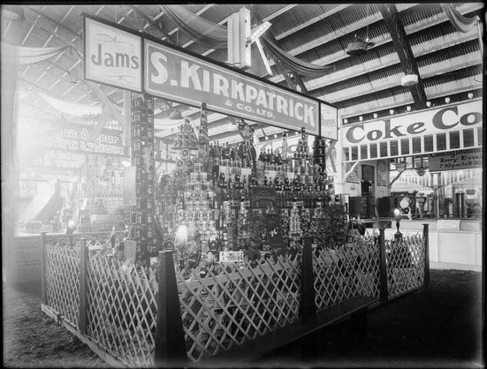 Black-and-white photo: Display stand for S. Kirkpatrick & Co., Ltd, makers of jams and preserves. 1890s–1930s. Photographer, Samuel Heath Head. Description: An indoor display of tinned and bottled produce several shelves high, surrounded by a low trellis fence. A long sign with the company name and the words “Jams“ (at left) and “Preserves” (at right) is mounted above the stacked items. Citation: Head, Samuel Heath, 1868-1948: Negatives. Ref: 1/1-007212-G. Alexander Turnbull Library, Wellington, New Zealand. https://natlib.govt.nz/records/23197778
