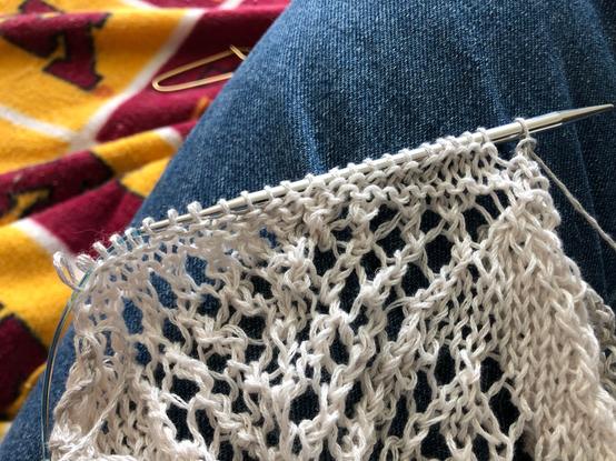 A photograph centred on a portion of a knitting project, the last stitch pattern repeat to be bound off, resting on the denim-clad thigh of the knitter. In the out-of-focus background is a stitch holder on a blanket of maroon and gold pattern.