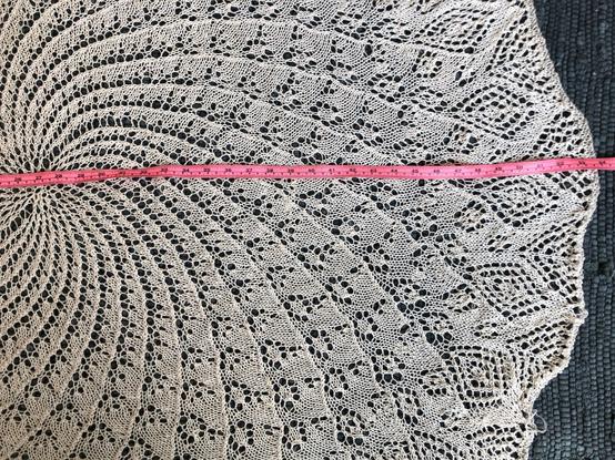 A portion of a knitted object, in off-white yarn laying on a dark blue woven rug, with a pink measuring tape across the centre and on to the far edge. Zooming in on that edge shows a measurement of about 136 cm / 53.5", while the centre is roughly 68.5 cm / 27".
