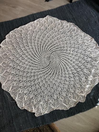 An image of the whole knitted object, in off-white yarn laying on a dark blue woven rug. The circular shawl awaits being washed and blocked using pins to bring out the 32 'points' at the end of each swirl. That step will enhance the visual geometry of the lace pattern.