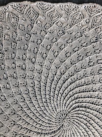 A portion of a knitted object, in off-white yarn laying on a dark blue woven rug, highlighting the lace pattern. It shows the progression from the centre toward the edge, and how each swirl has a series of 'bubbles' which start as a single hole and expands to a double diamond near the edge.