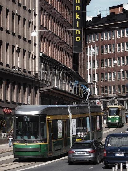 A street scene featuring a green tram line and a mix of vehicles, including a taxi. The background includes buildings with a visible sign for "Finnkino." The atmosphere is urban, with clear skies and pedestrians in view.