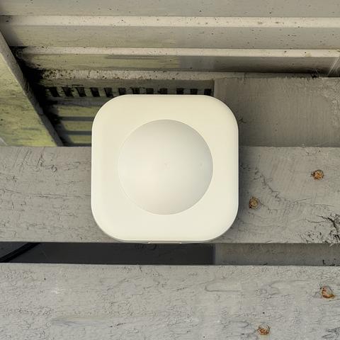 An IKEA Vallhorn motion sensor mounted on a fence. It’s a white square with rounded corners, and a small domed sensor in the middle.