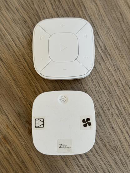Two IKEA remote controls. The one at the top is the new one, it’s a white rounded square with various buttons: “play” in the centre, plus and minus at the top and bottom, back and forward at top left and right, and one and two dots bottom left and right. 

Below that is our old remote. It’s the same shape but has only four buttons, light at the top, and then stickers for fan, sleep, and curtains going clockwise.