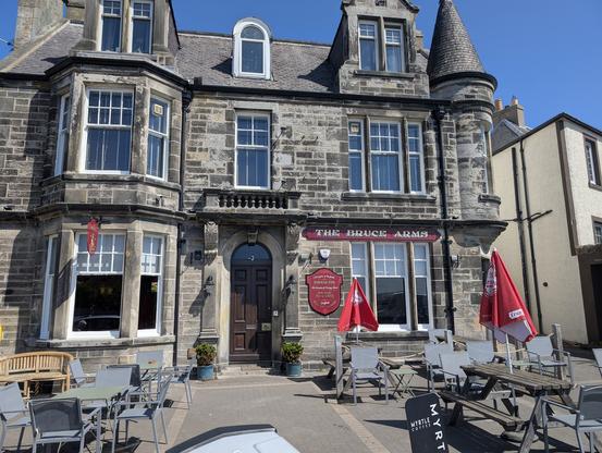 The image shows a three-story stone building with a classic architectural style, featuring a dark wooden door and large windows with white frames. The building is named "The Bruce Arms," as indicated by a red sign above the entrance. The facade includes a bay window on the left and a conical tower on the right, adding to its historical charm. The ground floor has several outdoor tables and chairs, some with red umbrellas, suggesting a cafe or pub setting. The sky is clear and blue, indicating a sunny day. The building is situated on a paved area with a few potted plants near the entrance. Adjacent to the building, there is a smaller structure with a different architectural style.