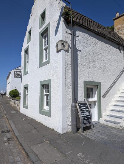 The image shows a white building with green window frames, located on a narrow street. The building has a sloped roof with dark tiles and a small chimney. There are several windows with white frames and green sills, and a small door with a green frame on the right side, leading to a set of white stairs. A signboard on the sidewalk reads "Sundial Cafe" and another sign indicates the "Toilets Entrance Behind Stairs." The building's exterior is textured, and there is a small bush near the entrance. The sky is clear and blue, suggesting a sunny day. The street is paved with cobblestones, and there is a street lamp visible. The building appears to be a cafe or small restaurant, as indicated by the signboards.