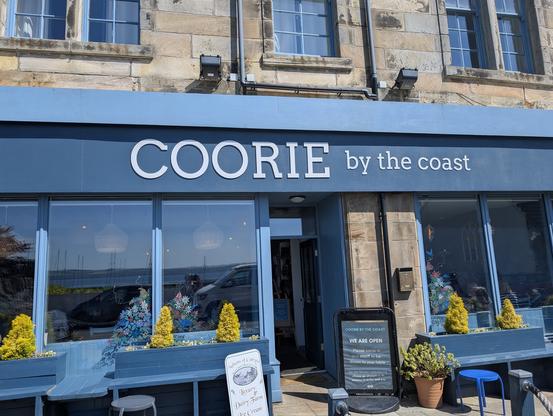 The image shows the exterior of a shop named "COORIE by the coast." The building has a stone facade with large windows and a blue awning above the entrance. The shop's name is prominently displayed in white capital letters on the blue awning. The windows are framed in blue, and there are decorative plants with yellow foliage in the window boxes. Inside, colorful floral arrangements are visible through the glass. A sign in front of the entrance reads "WE ARE OPEN" and mentions "Luxury Dairy Farm Ice Cream." The shop appears to be open for business, with a welcoming atmosphere. The overall color scheme is blue and white, with natural stone tones from the building.