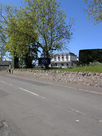 The image depicts a serene scene featuring a two-story white building with a dark roof, identified as "The Inn at Charlestown," as indicated by the sign in the foreground. The building is surrounded by lush greenery, including a large tree with vibrant green leaves and a well-maintained hedge. A stone wall runs along the front of the property, separating it from a quiet road with visible lane markings. The sky is clear and blue, suggesting a sunny day. In the background, a smaller building with a light-colored facade is partially visible. The overall setting appears peaceful and inviting, with the inn prominently displayed against the backdrop of a sunny day.