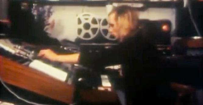 The image shows a person operating a large sound mixing console in a recording studio. The individual is focused, with one hand adjusting the controls. Behind them, there are two large reel-to-reel tape machines, their distinctive circular spools visible. The surroundings are dimly lit, with various audio equipment scattered around, creating a sense of a busy, technical workspace. The person has light-colored hair and is wearing dark clothing.