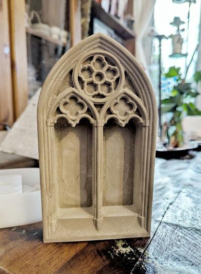 Dark table. On the table a small cathedral window made out of concrete. It is in a gothic style with 2 deep niches and each with one quatrefoil flowers under a bigger, solitaire, quintefoile styled element.