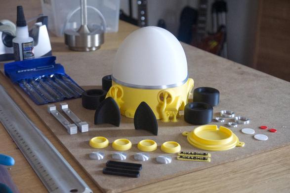 Table shot with parts of a 3D printyed model on display. These are parts of the pikachibi cat car night light. We see the main body with the print supports still attached in the middle. All the other parts like the tires, wheelcaps, lisence plates, cat ears, front and tail lights are grouped around the main body. Some tools are laying on the table as well.