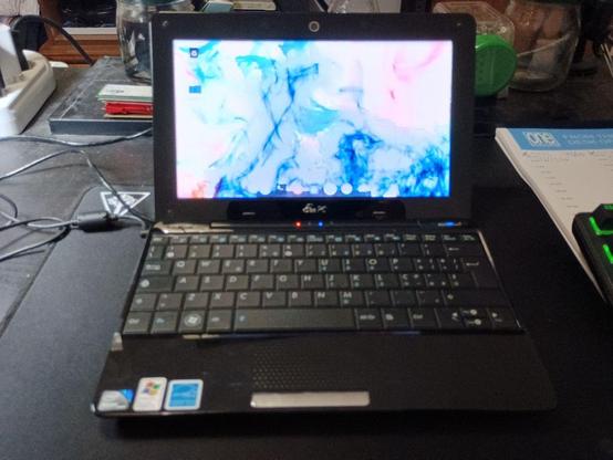 L'immagine mostra un laptop aperto su una superficie scura, probabilmente una scrivania. Il laptop è di colore nero con una tastiera nera e un display che mostra uno sfondo astratto con colori blu e rosa. Sulla tastiera sono visibili diverse etichette, tra cui una con il logo di Windows. Il laptop è collegato a un cavo, che si estende verso sinistra. Sullo sfondo, si possono vedere vari oggetti, tra cui un libro, un contenitore di vetro e un foglio con scritte. A destra del laptop, si intravede un'altra tastiera con luci verdi. L'immagine è leggermente sfocata, ma i dettagli principali sono chiari.