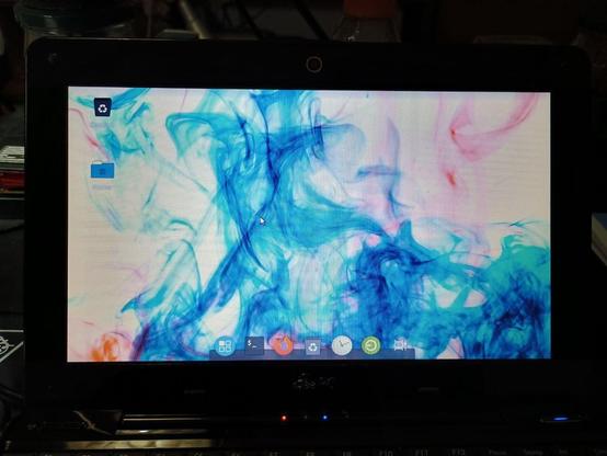 L'immagine mostra un laptop con lo schermo acceso, visualizzando un desktop con uno sfondo astratto di inchiostro che si mescola in colori blu, rosa e bianco. Sullo schermo sono visibili alcuni iconi, tra cui un file di riciclo e un file di cartella, situati nell'angolo in alto a sinistra. In basso, sulla barra delle applicazioni, sono presenti icone per vari programmi, tra cui un pulsante di menu, un simbolo di dollaro, un pulsante di ricarica, un orologio e un pulsante di configurazione. Il laptop è posizionato su una superficie scura, con un'atmosfera generale di un ambiente informale, probabilmente un ufficio o una stanza personale.