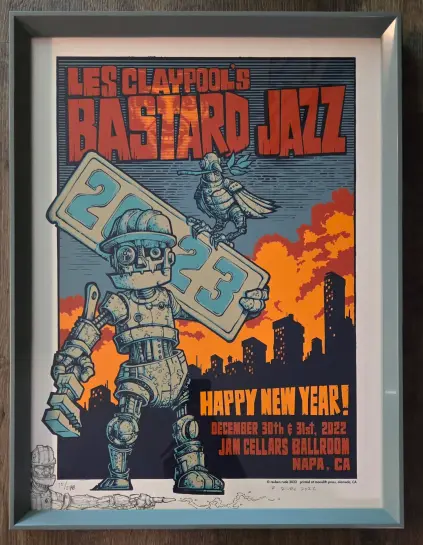 The image is a framed poster for a music event featuring "Les Claypool's Bastard Jazz." The poster has a vibrant design with a robot holding a sign that reads "2023," symbolizing the New Year. The robot is depicted in a mechanical style with a hard hat, and a bird is perched on its shoulder. The background shows a city skyline with a sunset sky in shades of orange and red. The text "Happy New Year!" is prominently displayed in bold orange letters, along with the event dates "December 30th & 31st, 2022" and the venue "Jam Cellars Ballroom, Napa, CA." The poster is signed by the artist, Reuben Rude, and includes a print number "15/278." The overall color scheme is a mix of blue, orange, and black, creating a striking visual contrast.