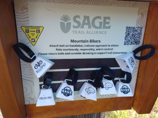 This is a sign at a trailhead for bikes at Elings Park.  The border margins depict elevation contour lines of the type seen on topographic maps.  The top middle shows a logo that resembes a plant before the "SAGE Trail Alliance" heading.
Below, center aligned text says: 
Mountain Bikers
Attach bell on handlebar, indicate approach to others
Rid courteously, responsibly and in control
Please return bells and consider donating to support trail sponsorship.
On the left there is a yellow and black graphic of a upside down triangular sign 
explaining trail courtesy: At the upper left corner, a bike rider graphic, an arrow point to the right corner graphic of a hiker.  Below that, text says "Yield To"
Two arrows point to a horseback rider at the bottom corner from the bike rider and hiker graphics.  On the right, there is a QR code, for a donation link. 
The sign has 3 black magnetized strips, to which cling 7 small cow-bell shaped bells with Velcro straps.  The bells display logos for either VeloPro Cyclery, SAGE trail alliance, or a drawing of a hand gripping a handle bar, with the letters G R I T written on 4 finger segments facing forward.