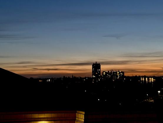 The sky changes colour from blue to orange on the horizon, it is beginnig to get dark and there are lights in a silhouetted building in the distance.