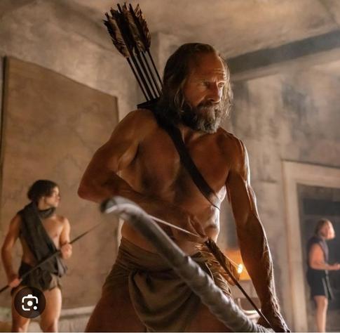 A VERY cut (very muscular) Ralph Fiennes tanned, long haired, with scruffy beard and mustache. Stands barechested with a loincloth and a bow and arrow. Behind him are young men similarly clad.