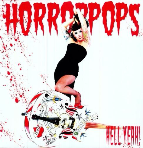 Photo of Patricia Day from HorrorPops standing with one knee bent on a white standup bass. Bass is lying on the ground, it is adorned with traditional style tattoo art. Patricia is wearing a black slinky vintage dress and red heels, heavily tattooed arms. A white background is splattered with blood and the word Horror Pops is across the top of the imagine in red spooky letters.