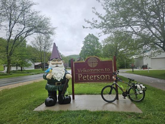 A bicycle with a white bag leans against  a brown sign that says in gold letets, "Velkommen to Peterson". The sign is on a square piece of concrete surrounded by green grass. Next to the sign is a large gnome with a pointy purple hat, white beard, green tunic, black belt with a gold buckle, and black boots. A blue bird with a yellow chest is on its right hand.
A tree is behind it. An asphalt trail is on the right, a street is on the left, a few buildings along it.