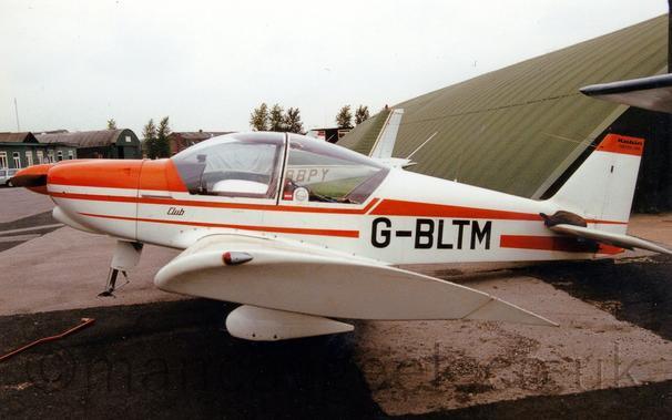 Side view of a single-engined light aircraft parked facing to the left, with it's tail weighed down so it's nose, minus the nose-wheel, is raised in the air.
The plane is mostly white, with bright orange stripes running along the body, and the black registration "G-BLTM" on the lower rear fuselage.
There is a green hangar in the background on the right, with more green buildings in the distance on the right, under flat grey skies.