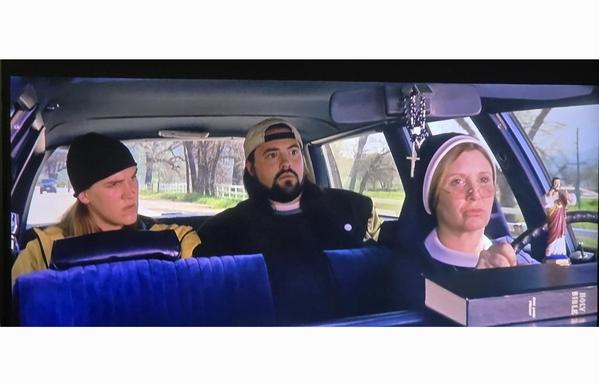 Jason Mewes, Kevin Smith and Carrie Fisher dressed as a nun in Jay and Silent Bob Strike Back. They're in the car back seat looking surprised. She's driving with a smiling dashboard Jesus with two thumbs up.