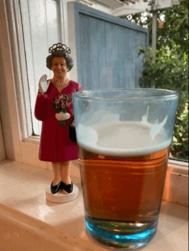 A blue glass of golden-red liquid with white foam sits in the su, in a white painted windowsill. Behind the glass, a small statue of Queen Elizabeth smiles and waves at the camera.