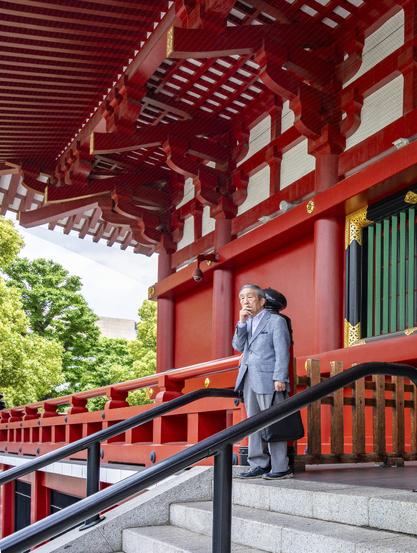 A man in blue grey suit is standing at the top of stairs leading to the bright red Senso Ji temple, In Asakusa, in Tokyo, Japan. He holds one hand on his mouth and seems to be hesitating. He is holding a work case in the other hand.