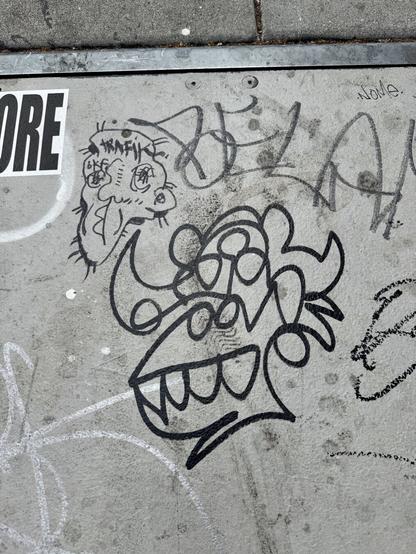 Graffitin on the sidewalk. The big piece in the center looks like a gorilla's head, but with a third eye on the forehead. It is drawn in black ink in one single conintious line. This is Plasmo, the graffiti character drawn by an artist that is also named Plasmo. There is another cool graffiti tag to the left of this piece which is a drawing of a man's head by an artist named Trafik.