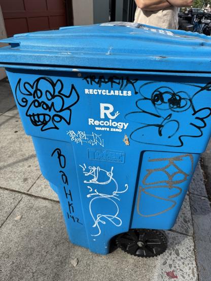 Tags by Plasmo and Trafik on a blue trashcan. Plasmo drew his gorilla character and Trafik drew a man's head. There are many other tags on the can.